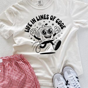 Op de afbeelding: Crèmekleurig T-shirt met de tekst "LIFE IN LINES OF CODE" en een cartoonafbeelding van een retro televisiefiguur. Rood-witte geruite shorts en grijze sneakers met zwarte strepen zijn ook zichtbaar.