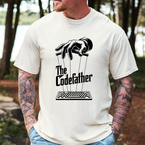 Op de afbeelding: Crèmekleurig T-shirt met een zwart-witte afbeelding van een hand die een toetsenbord met touwtjes bestuurt. De tekst "The Codefather" staat eronder. Het shirt heeft korte mouwen.