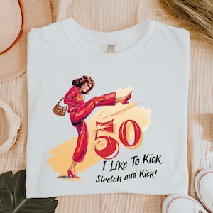 Puede incluir: Camiseta blanca con una ilustración roja y blanca de una mujer pateando un gran número rojo 50. El texto "I Like To Kick. Stretch and Kick!" está impreso debajo de la ilustración.