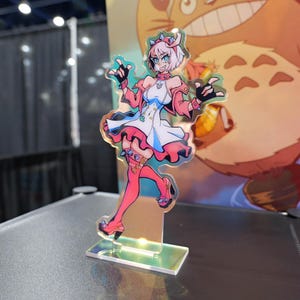 Könnte beinhalten: Ein farbenfrohes Acryl-Standee mit einer Cartoon-Figur mit rosa Haaren, einem weißen Kleid und rosa Akzenten. Die Figur posiert mit erhobenen Händen, und das Standee hat einen holografischen Effekt. Der Hintergrund ist unscharf.