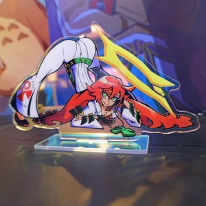 May include: A clear acrylic stand featuring a stylized anime character with red hair, green eyes, and a white outfit. The character is in a dynamic pose with a yellow and blue design element. The base is rectangular and clear.