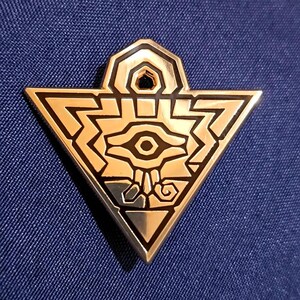 Puede incluir: Un broche triangular de metal dorado con un símbolo de ojo estilizado en el centro. El diseño incluye líneas geométricas intrincadas y detalles en esmalte negro, con un pequeño lazo circular en la parte superior para sujeción.