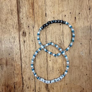 May include: Two beaded bracelets with the words "PIXIE DUST". The bracelets are made of white, blue, and clear beads. The letters are black and white. The bracelets are on a wooden surface.