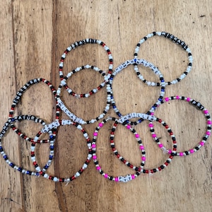 May include: A collection of ten circular beaded bracelets featuring white letter beads spelling out song titles and names including BEAT IT, MICHAEL, HUMAN NATURE, THRILLER, and PYT, interspersed with small black, red, white, blue, and pink plastic beads.