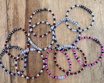 May include: A collection of ten circular beaded bracelets featuring white letter beads spelling out song titles and names including BEAT IT, MICHAEL, HUMAN NATURE, THRILLER, and PYT, interspersed with small black, red, white, blue, and pink plastic beads.