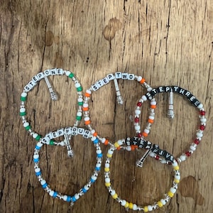 May include: Five circular beaded bracelets featuring white letter beads spelling out STEP ONE, STEP TWO, STEP THREE, STEP FOUR, and STEP FIVE. Each bracelet includes a small silver-toned microphone charm and colorful accent beads in green, orange, red, blue, and yellow.