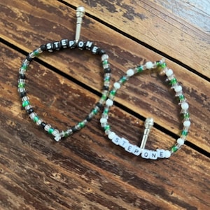 May include: Two beaded bracelets with the text "STEP ONE". One bracelet is black, green, and clear beads, the other is white, green, and clear beads. Both bracelets have a silver-colored metal clasp.