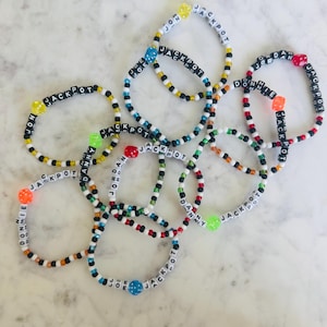 May include: A collection of beaded bracelets with black and white letter beads spelling out names and the word "JACKPOT". The bracelets feature colorful beads and dice-shaped accents. The bracelets are on a white marble surface.
