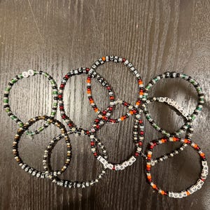 May include: A collection of handmade beaded bracelets featuring black, red, orange, and green beads. Several bracelets include white letter beads spelling out ZACH BRYAN, REVIVAL, BURN, and CONDEMNED, along with number beads displaying 28.