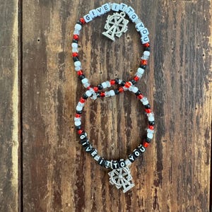 May include: Two beaded bracelets with the text "GIVE IT TO YOU" in white letter beads. The bracelets have red, black, and white beads, and a silver charm of a Ferris wheel. The bracelets are on a wooden surface.