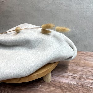 Könnte beinhalten: Ein hellgrauer Fleece-Stoff, der über einem Holzständer mit natürlicher Oberfläche drapiert ist. Der Stoff ist weich und texturiert, und der Ständer hat drei Beine. Ein getrockneter Grashalm liegt auf dem Stoff.