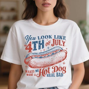 May include: White t-shirt with a graphic design featuring a hot dog and text that reads "You look like the 4th of July makes me want a hot dog real bad" in red and blue.