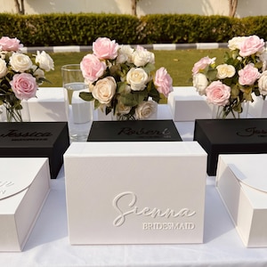 Puede incluir: Una caja de regalo rectangular blanca con el nombre Sienna y la palabra BRIDESMAID en relieve en la parte frontal. La caja se muestra junto a otras cajas negras y blancas, cada una con nombres y roles personalizados, acentuadas por ramos de rosas rosas y blancas.