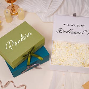 Pu&ograve; includere: Una pila di due scatole regalo rettangolari color verde oliva e verde acqua, ciascuna legata con un nastro. Una terza scatola bianca &egrave; aperta, rivelando carta triturata all'interno e la scritta "WILL YOU BE MY Bridesmaid ?" sul coperchio.