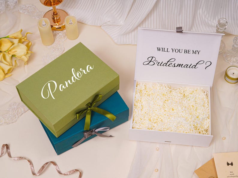 Pu&ograve; includere: Una pila di due scatole regalo rettangolari color verde oliva e verde acqua, ciascuna legata con un nastro. Una terza scatola bianca &egrave; aperta, rivelando carta triturata all'interno e la scritta "WILL YOU BE MY Bridesmaid ?" sul coperchio.