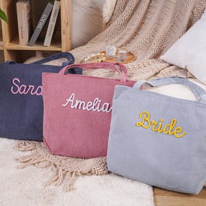 May include: Three corduroy tote bags in navy, pink, and light blue. Each bag has a handle and embroidered text: "Sara", "Amelia", and "Bride". The bags are on a white rug with a neutral background.