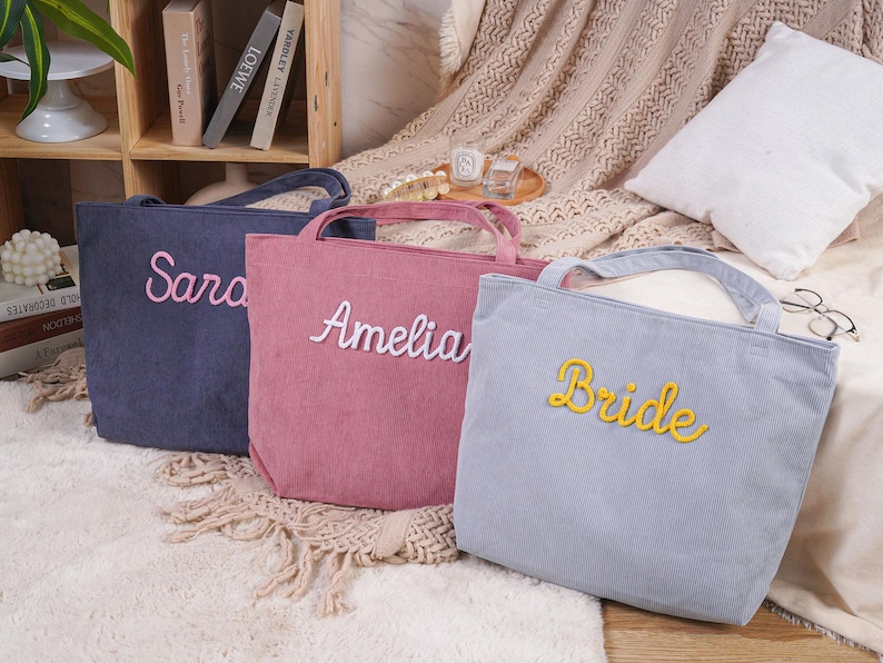 May include: Three corduroy tote bags in navy, pink, and light blue. Each bag has a handle and embroidered text: "Sara", "Amelia", and "Bride". The bags are on a white rug with a neutral background.