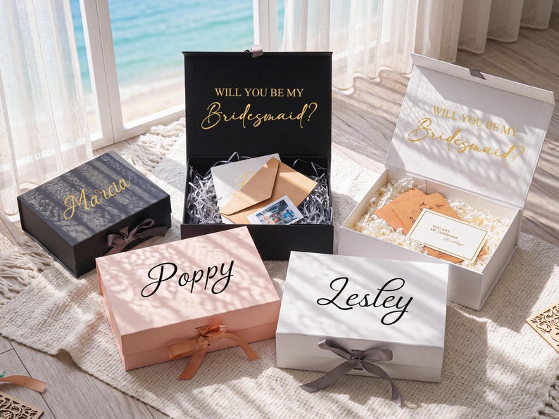 Pu&ograve; includere: Una collezione di scatole regalo rettangolari nere, bianche e rosa chiaro, dotate di chiusure a nastro e nomi personalizzati in corsivo. Due scatole aperte mostrano il testo Will You Be My Bridesmaid in lettere dorate sul coperchio interno.