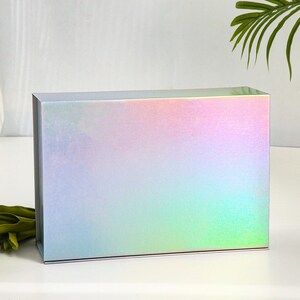 K&ouml;nnte beinhalten: Eine rechteckige Geschenkbox mit holografischem Finish, die einen Farbverlauf aus Pastellfarben wie Rosa, Blau und Gr&uuml;n zeigt. Die Box steht auf einer wei&szlig;en Oberfl&auml;che, mit gr&uuml;nen Stielen und wei&szlig;en Blumen im Vordergrund.