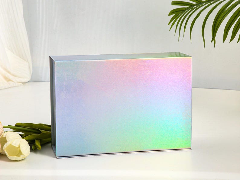 K&ouml;nnte beinhalten: Eine rechteckige Geschenkbox mit holografischem Finish, die einen Farbverlauf aus Pastellfarben wie Rosa, Blau und Gr&uuml;n zeigt. Die Box steht auf einer wei&szlig;en Oberfl&auml;che, mit gr&uuml;nen Stielen und wei&szlig;en Blumen im Vordergrund.