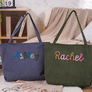 May include: Two corduroy tote bags, one blue and one green, with embroidered names. The blue bag says "Ashley" in blue, green, and white thread. The green bag says "Rachel" in tan and pink thread. Both have long handles.