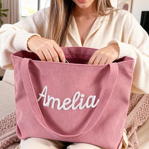 May include: A dusty rose corduroy tote bag with the name "Amelia" embroidered in white script. The bag has long handles and a zippered top. The image shows the bag being opened.
