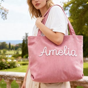 May include: A pink corduroy tote bag with the name "Amelia" embroidered in white script. The bag has a rectangular shape with two handles. The background shows a person wearing a white shirt and a scenic outdoor view.