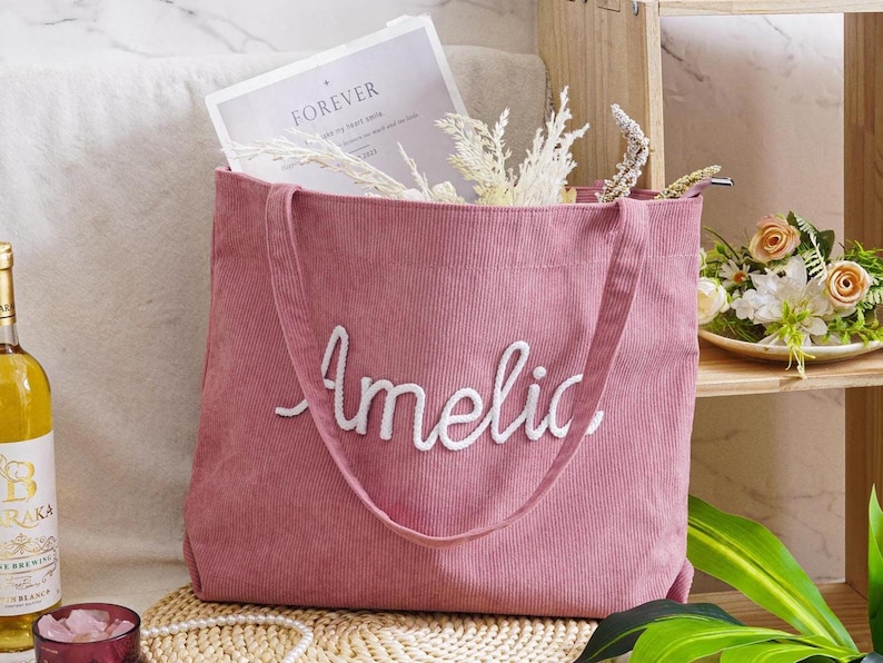 May include: A dusty rose corduroy tote bag with the name "Amelie" embroidered in white script. The bag has long handles and is filled with a magazine and dried flowers. A bottle of wine and a small dish are in the background.