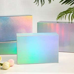 K&ouml;nnte beinhalten: Mehrere rechteckige Geschenkboxen mit schillernden, farbwechselnden Oberfl&auml;chen. Die Boxen weisen eine Mischung aus Rosa, Blau und Gr&uuml;n auf. Sie stehen auf einer wei&szlig;en Oberfl&auml;che, mit Blumen und Gr&uuml;nzeug im Vordergrund.