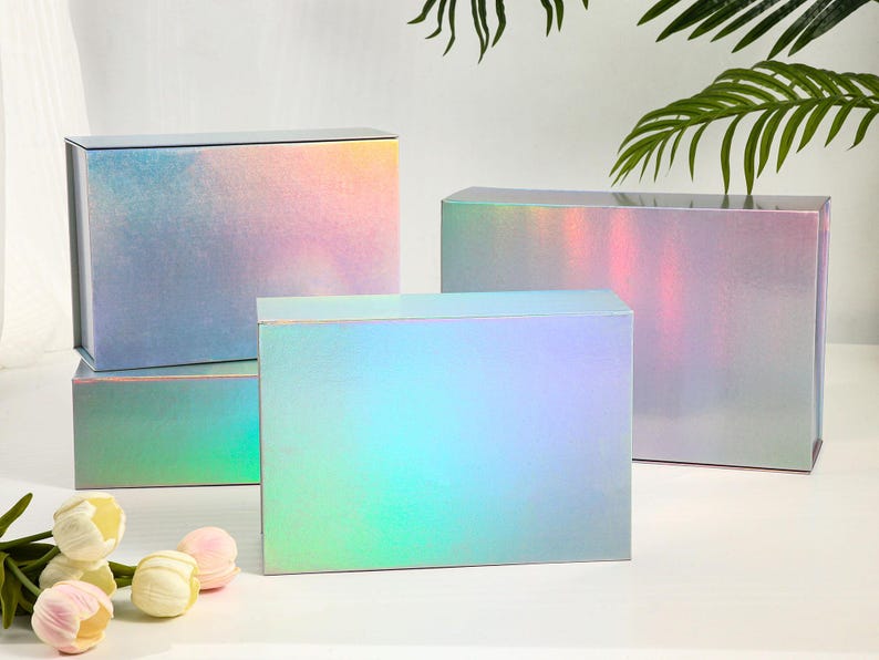 K&ouml;nnte beinhalten: Mehrere rechteckige Geschenkboxen mit schillernden, farbwechselnden Oberfl&auml;chen. Die Boxen weisen eine Mischung aus Rosa, Blau und Gr&uuml;n auf. Sie stehen auf einer wei&szlig;en Oberfl&auml;che, mit Blumen und Gr&uuml;nzeug im Vordergrund.