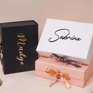 Pu&ograve; includere: Una scatola regalo nera verticale con il nome Madge in corsivo dorato, una scatola bianca orizzontale con il nome Sabrina in corsivo nero e una scatola rosa chiaro orizzontale, tutte dotate di chiusure a nastro e coperchi magnetici.
