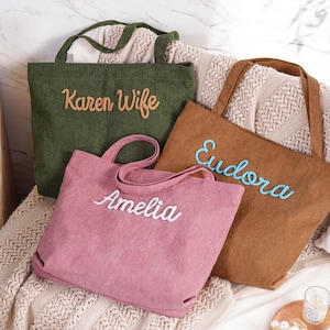 May include: Three corduroy tote bags in olive green, brown, and pink, each embroidered with a name in a contrasting color. The bags have sturdy handles and a simple, rectangular design, perfect for carrying items.
