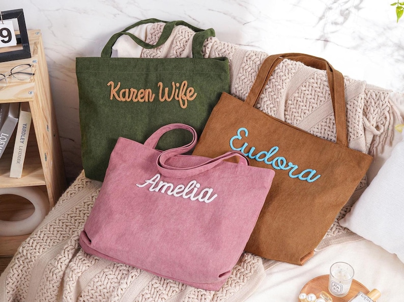 May include: Three corduroy tote bags in olive green, brown, and pink, each embroidered with a name in a contrasting color. The bags have sturdy handles and a simple, rectangular design, perfect for carrying items.
