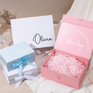 Pu&ograve; includere: Una collezione di scatole regalo in vari colori, tra cui bianco, azzurro e rosa. Le scatole sono personalizzate con nomi e frasi, alcune con nastri. La scatola rosa &egrave; aperta, riempita di carta sminuzzata, e ha il testo "Will you be my Bridesmaid?" e un nome.