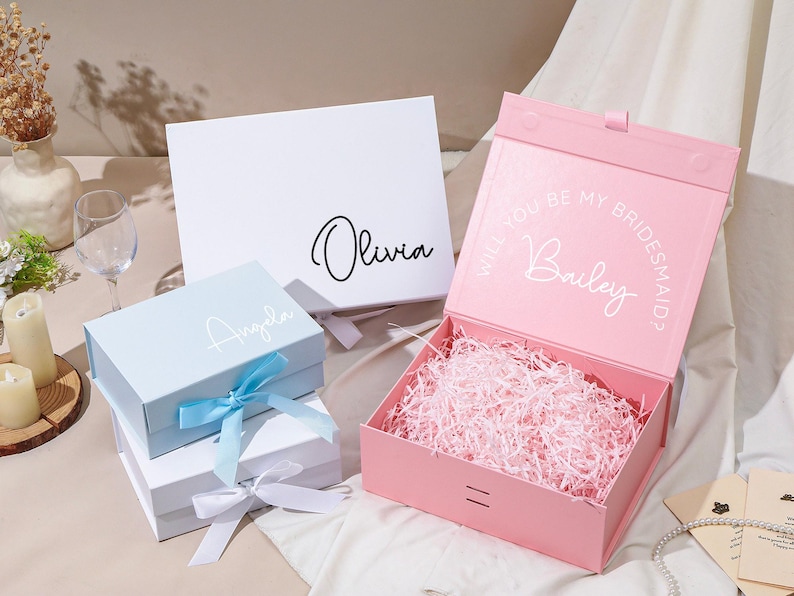 Pu&ograve; includere: Una collezione di scatole regalo in vari colori, tra cui bianco, azzurro e rosa. Le scatole sono personalizzate con nomi e frasi, alcune con nastri. La scatola rosa &egrave; aperta, riempita di carta sminuzzata, e ha il testo "Will you be my Bridesmaid?" e un nome.