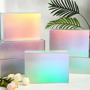 K&ouml;nnte beinhalten: Mehrere rechteckige Geschenkboxen mit schillernden, farbwechselnden Oberfl&auml;chen. Die Boxen sind gestapelt und einzeln angeordnet. Der Farbverlauf wechselt von Gelb &uuml;ber Rosa zu Blau. Einige k&uuml;nstliche Blumen befinden sich im Vordergrund.