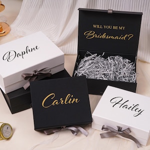 Pu&ograve; includere: Una collezione di scatole regalo rettangolari bianche e nere, dotate di chiusure magnetiche e nastri grigi. Le scatole sono personalizzate con nomi in corsivo nero o dorato. Una scatola nera aperta mostra il testo Will You Be My Bridesmaid?