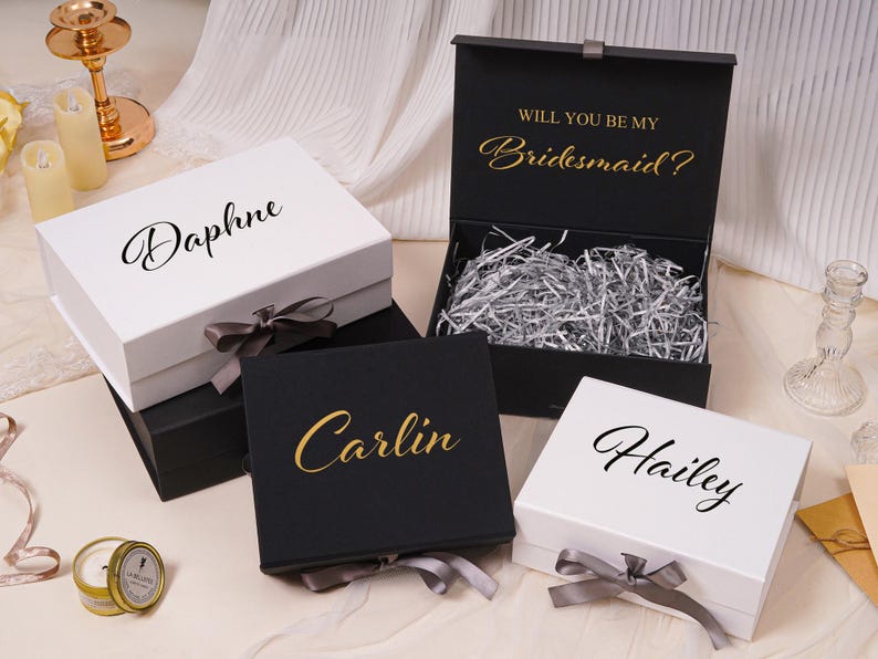 Pu&ograve; includere: Una collezione di scatole regalo rettangolari bianche e nere, dotate di chiusure magnetiche e nastri grigi. Le scatole sono personalizzate con nomi in corsivo nero o dorato. Una scatola nera aperta mostra il testo Will You Be My Bridesmaid?