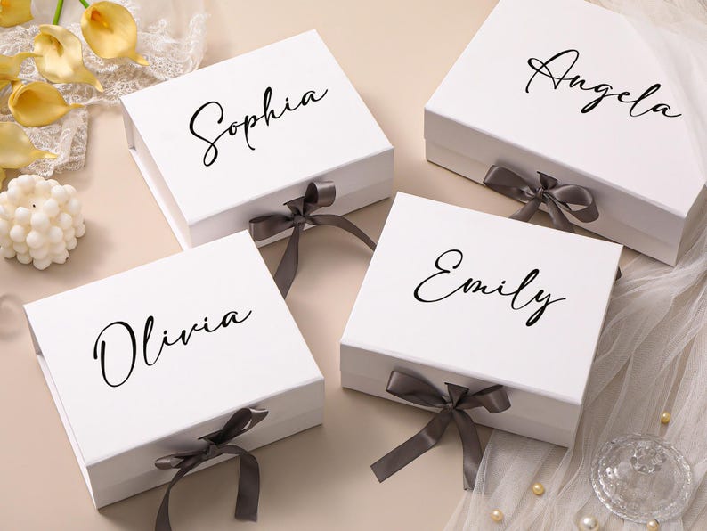 Pu&ograve; includere: Quattro scatole regalo rettangolari bianche con chiusure magnetiche e nastri grigio scuro. Ogni scatola presenta un nome personalizzato stampato in carattere corsivo nero sul coperchio, ovvero Sophia, Angela, Olivia ed Emily, disposte in gruppo.
