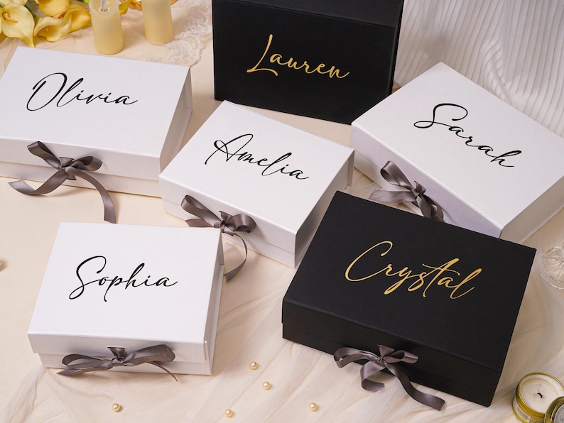 Pu&ograve; includere: Una collezione di scatole regalo rettangolari in bianco e nero, ognuna con un nome personalizzato in carattere corsivo. Le scatole bianche hanno testo nero e nastri grigi, mentre quelle nere hanno testo dorato e nastri grigi.