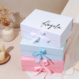 Pu&ograve; includere: Tre scatole regalo impilate. La scatola superiore &egrave; bianca con il nome "Angela" in corsivo nero. La scatola centrale &egrave; azzurra e quella inferiore &egrave; rosa. Ogni scatola ha un nastro abbinato legato con un fiocco.