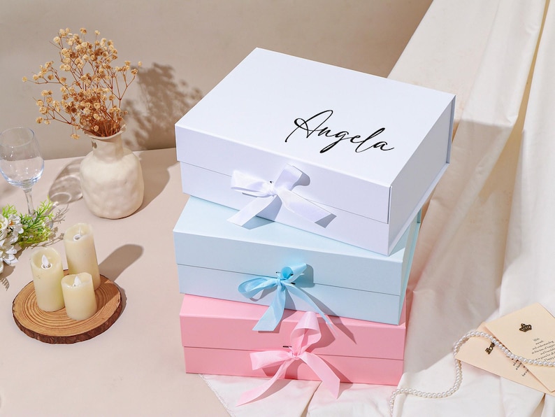 Pu&ograve; includere: Tre scatole regalo impilate. La scatola superiore &egrave; bianca con il nome "Angela" in corsivo nero. La scatola centrale &egrave; azzurra e quella inferiore &egrave; rosa. Ogni scatola ha un nastro abbinato legato con un fiocco.