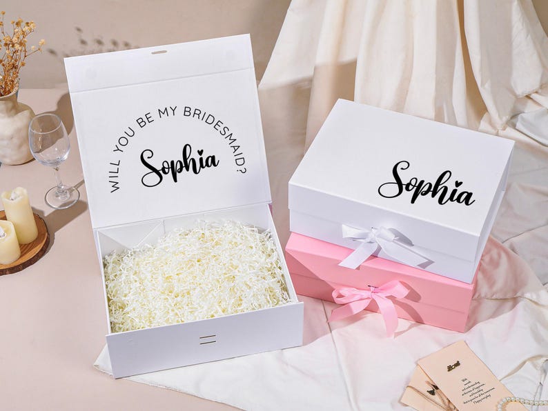 Pu&ograve; includere: Scatole regalo bianche, una aperta e una chiusa, con il nome "Sophia" in corsivo. La scatola aperta ha il testo "Vuoi essere la mia damigella d'onore?" ed &egrave; riempita di carta sminuzzata. Una scatola rosa &egrave; sotto la scatola bianca chiusa.