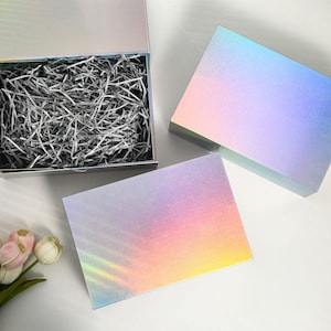 K&ouml;nnte beinhalten: Eine offene, silberne Geschenkbox mit zerknittertem Papier, neben zwei geschlossenen Boxen. Die Boxen haben eine holografische, irisierende Oberfl&auml;che, die Farben wie Rosa, Blau und Gelb reflektiert. Die Boxen stehen auf einer wei&szlig;en Oberfl&auml;che.