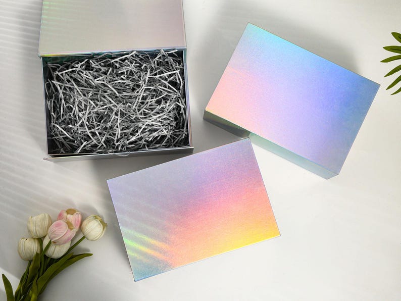 K&ouml;nnte beinhalten: Eine offene, silberne Geschenkbox mit zerknittertem Papier, neben zwei geschlossenen Boxen. Die Boxen haben eine holografische, irisierende Oberfl&auml;che, die Farben wie Rosa, Blau und Gelb reflektiert. Die Boxen stehen auf einer wei&szlig;en Oberfl&auml;che.