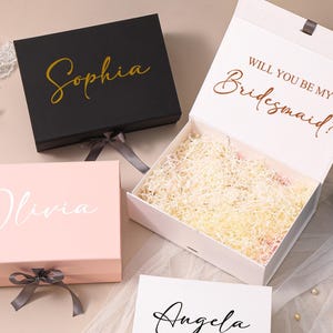 Pu&ograve; includere: Una collezione di scatole regalo rettangolari con chiusure magnetiche e nastri. Una scatola nera mostra il nome Sophia in oro, una scatola rosa mostra Olivia in bianco, e una scatola bianca aperta contiene carta sminuzzata con il testo WILL YOU BE MY BRIDESMAID? in rame.