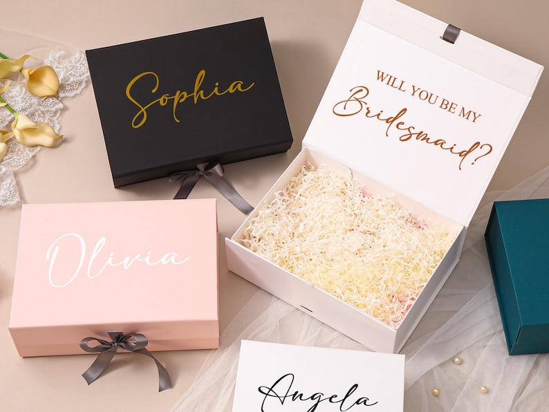 Pu&ograve; includere: Una collezione di scatole regalo rettangolari con chiusure magnetiche e nastri. Una scatola nera mostra il nome Sophia in oro, una scatola rosa mostra Olivia in bianco, e una scatola bianca aperta contiene carta sminuzzata con il testo WILL YOU BE MY BRIDESMAID? in rame.