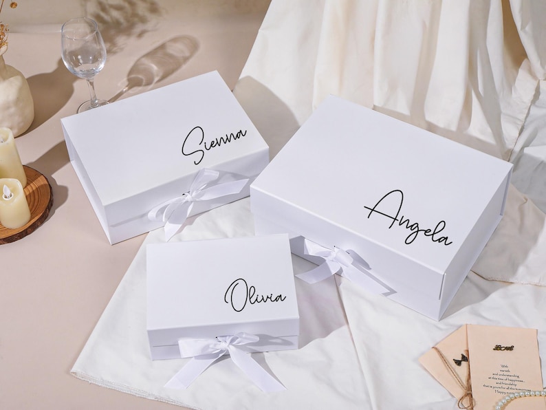 Pu&ograve; includere: Tre scatole regalo bianche con nomi in corsivo nero: Sienna, Angela e Olivia. Ogni scatola ha un nastro bianco legato a fiocco. Le scatole sono su una superficie bianca.