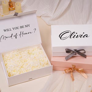 Pu&ograve; includere: Due scatole regalo rettangolari con chiusure magnetiche e nastri. Una scatola bianca &egrave; aperta, rivelando carta triturata all'interno e il testo WILL YOU BE MY Maid of Honor? stampato. Una seconda scatola bianca &egrave; chiusa con il nome Olivia stampato sopra.