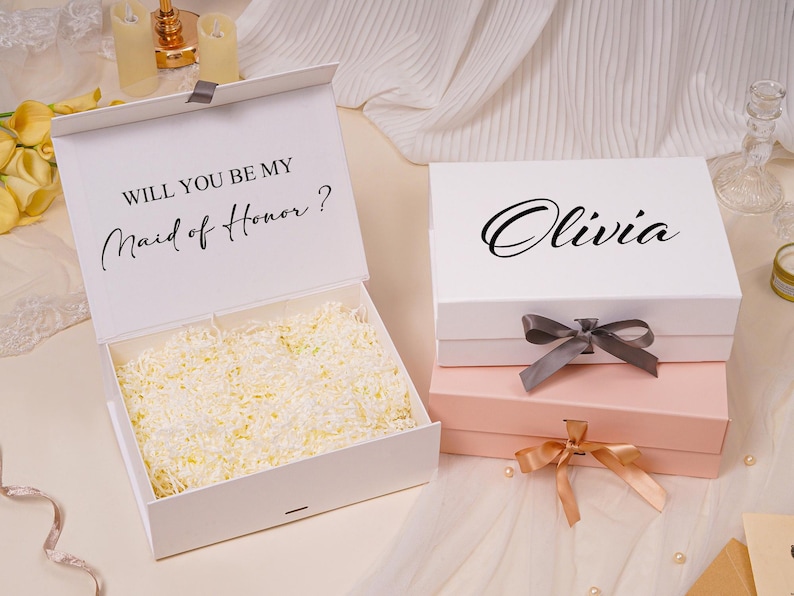 Pu&ograve; includere: Due scatole regalo rettangolari con chiusure magnetiche e nastri. Una scatola bianca &egrave; aperta, rivelando carta triturata all'interno e il testo WILL YOU BE MY Maid of Honor? stampato. Una seconda scatola bianca &egrave; chiusa con il nome Olivia stampato sopra.